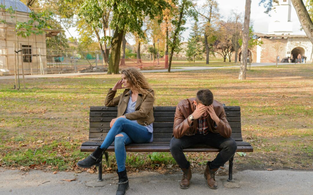 A couple sitting on the bench. One is looking away, while the other has his head in his hands.